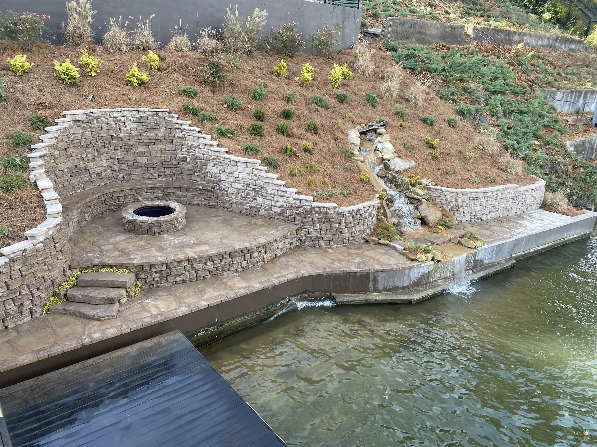 A curved stone wall at the lake’s edge with a fire pit and a spillway