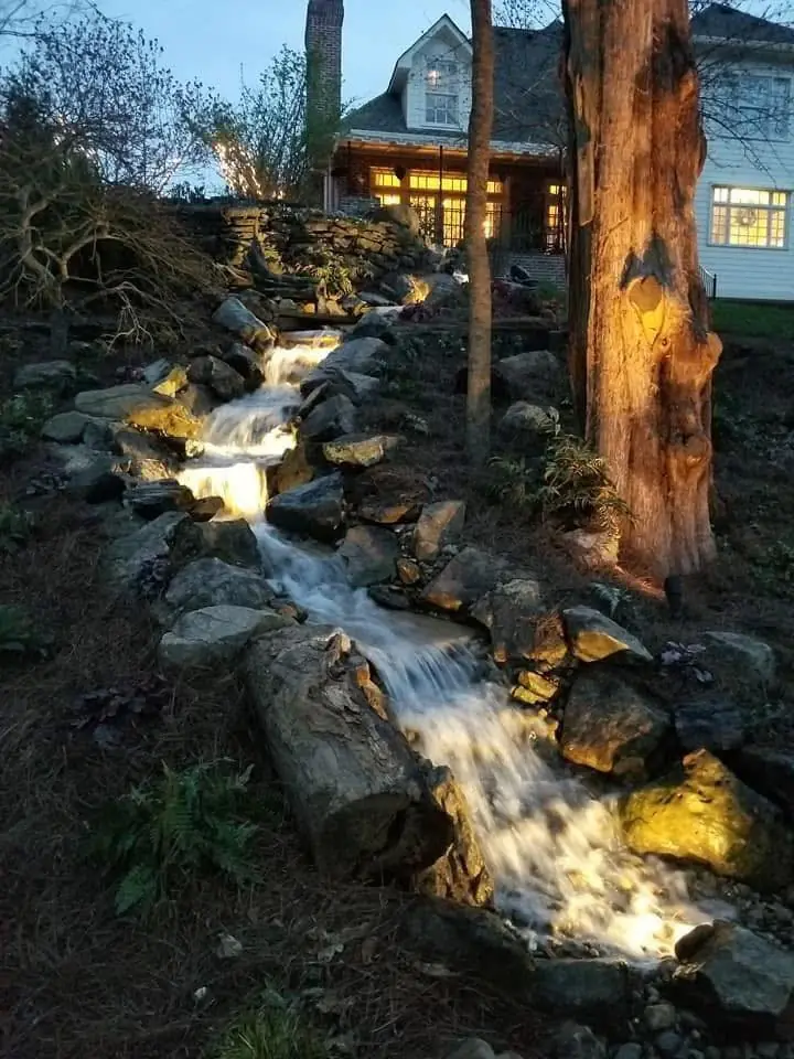 A lit stream falling past a tree trunk below a house at dusk
