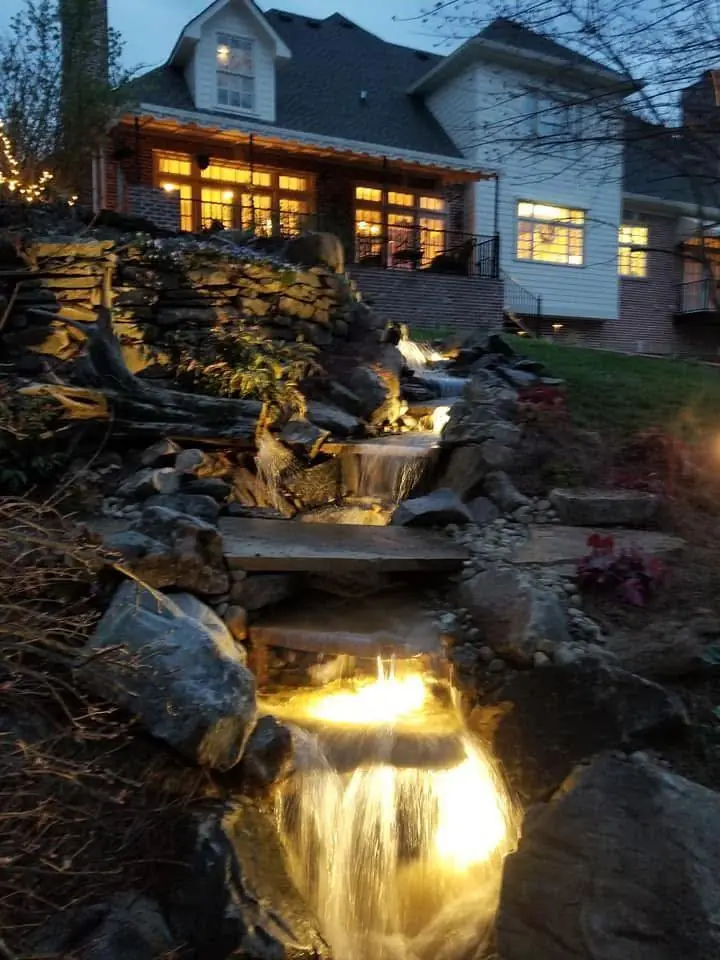 A lit waterfall below a house with its windows glowing at dusk