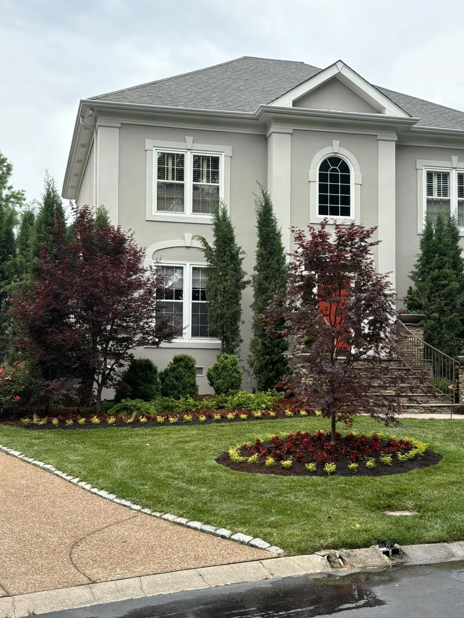A red maple ringed with flowers in front of a white house